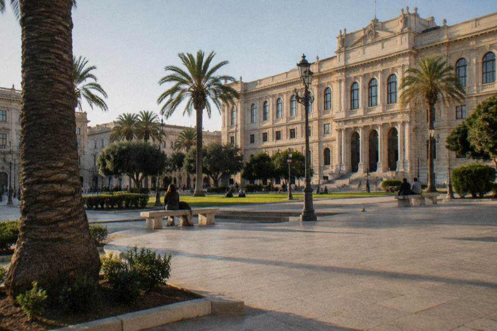 Broad city square in front of a monumental cream stone university building with palm trees, lawns and benches