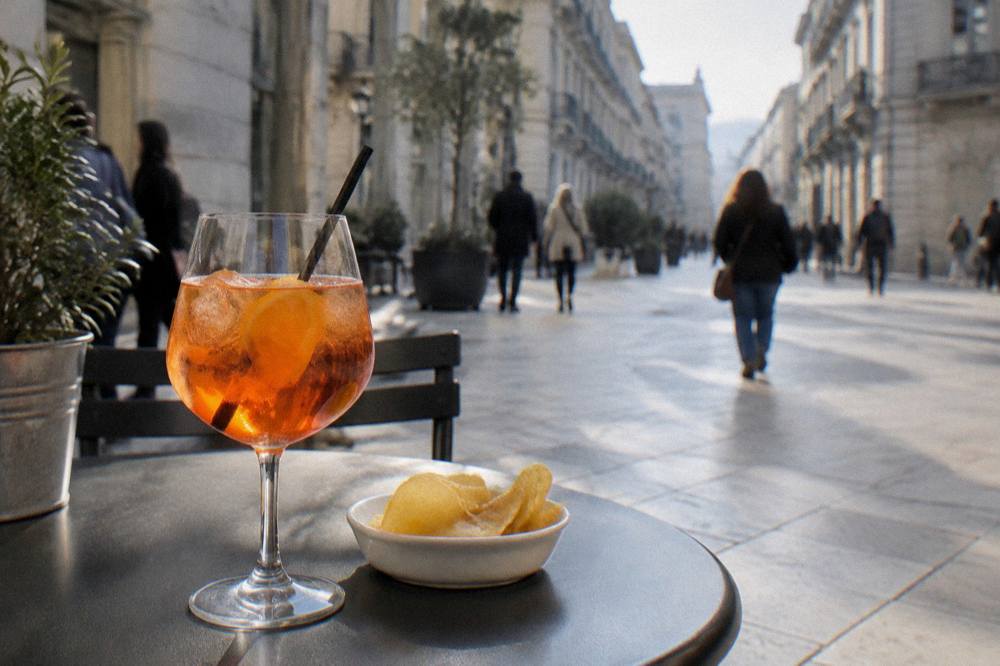 Pavement cafe table on a pedestrian street with an orange spritz and a small plate of crisps, passers-by blurred