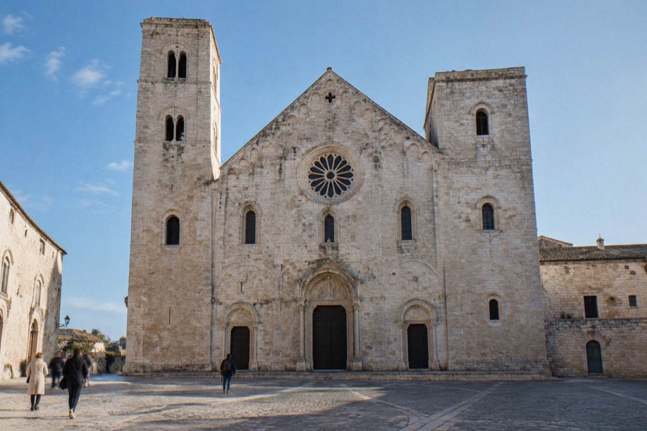 Romanesque church of pale limestone with a wide plain facade, two squat towers and a rose window above a paved square