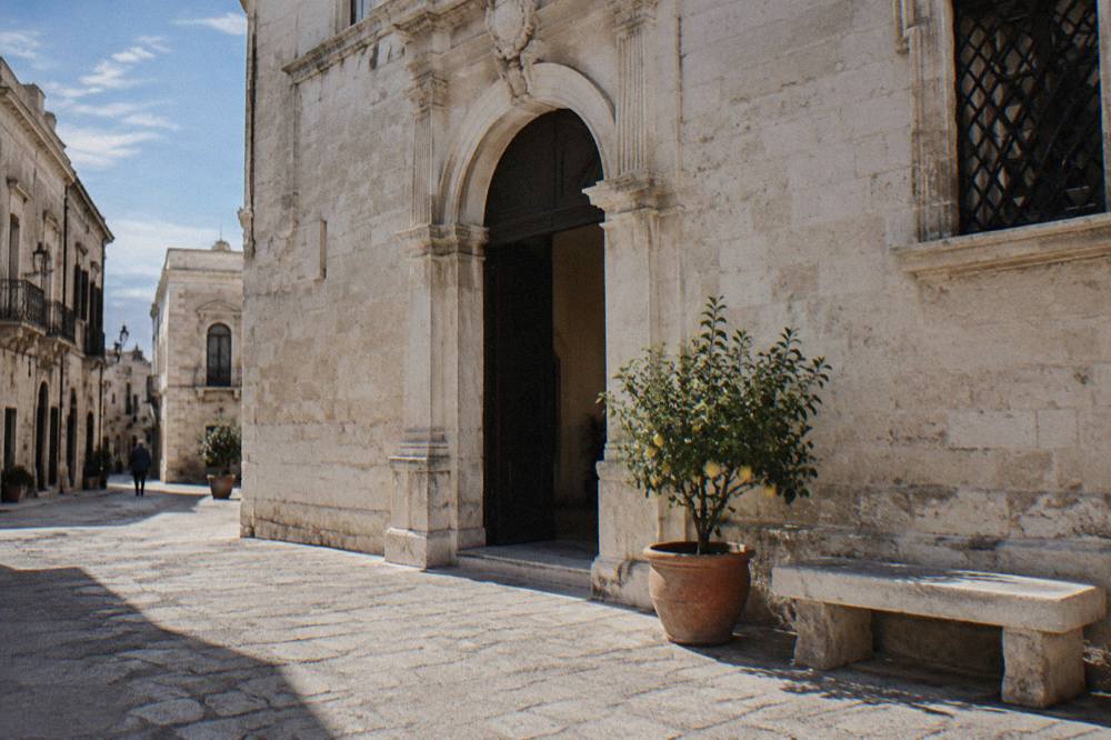 Pale stone palazzo with an arched portal on a quiet old-town street beside a stone bench and a potted lemon tree