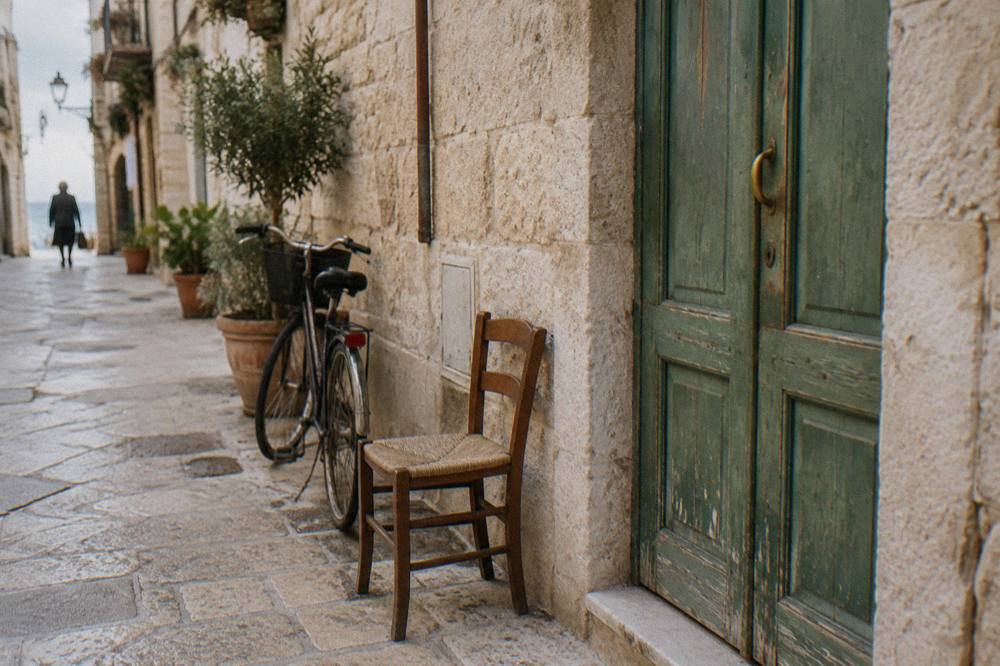 Faded green wooden door in a pale limestone wall with a chair and a bicycle beside it