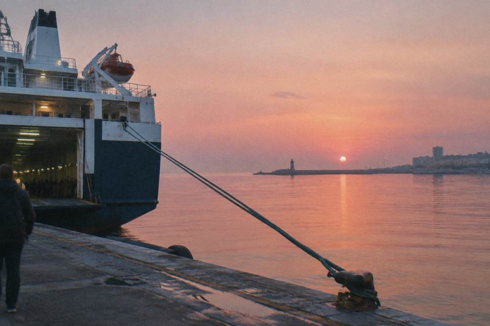 Stern of a ferry at a quay at sunrise with a pink sky, taut mooring ropes and still water