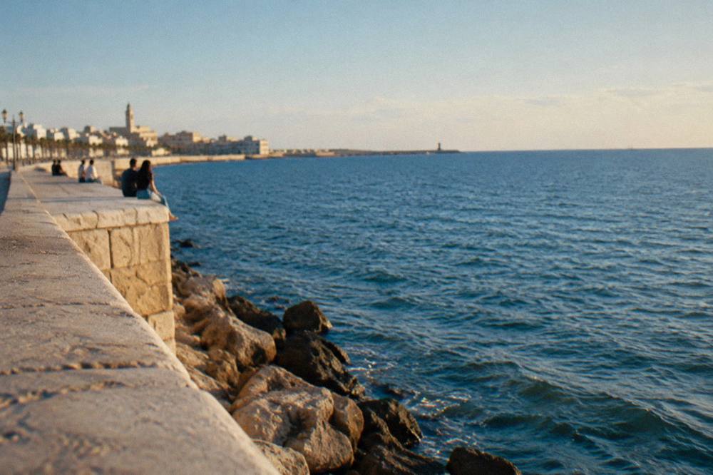 Distant people sitting on the broad top of a pale limestone sea wall above the open Adriatic in soft warm light