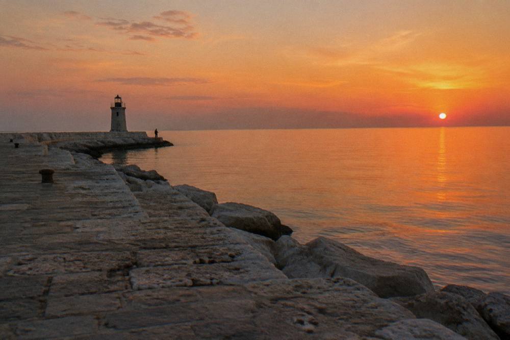 End of a harbour pier with a small lighthouse, glassy open Adriatic and a distant angler at sunset