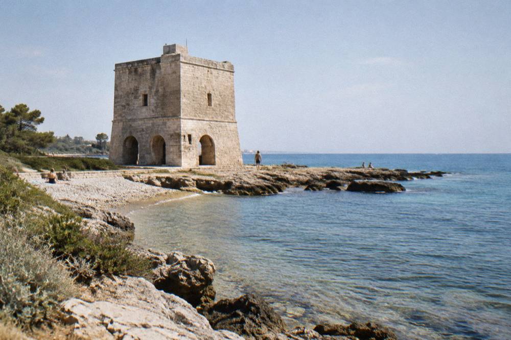 Squat square stone watchtower on a flat rocky shore beside a small pebble beach and low scrub