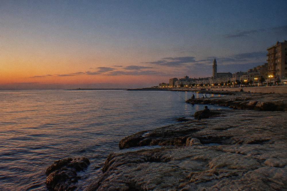 Seafront limestone rocks below a residential district with distant anglers and calm water in early evening