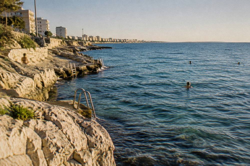 Small rocky bathing spot with ladders into the sea and a few distant swimmers