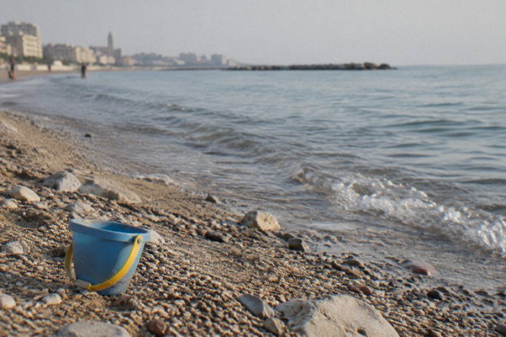 Pebbles and coarse sand at the water's edge with small clear waves and a plastic bucket left on the sand
