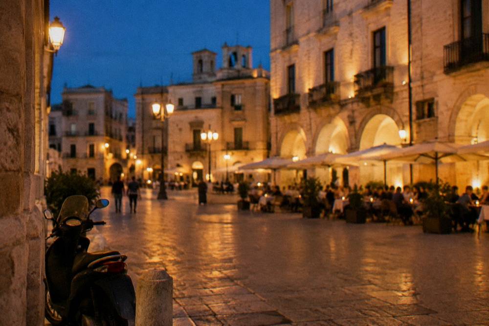 Evening piazza with warm light on cream walls, out-of-focus diners at tables and glowing market arches