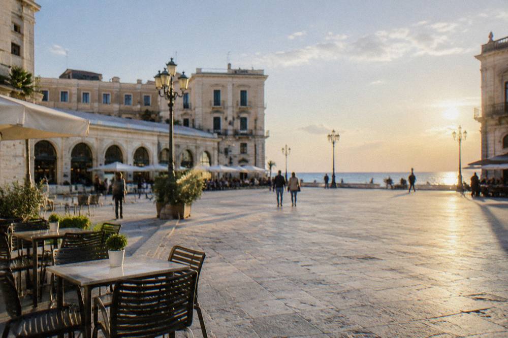 Broad open piazza beside a long low market hall with arched openings and outdoor cafe tables in bright morning