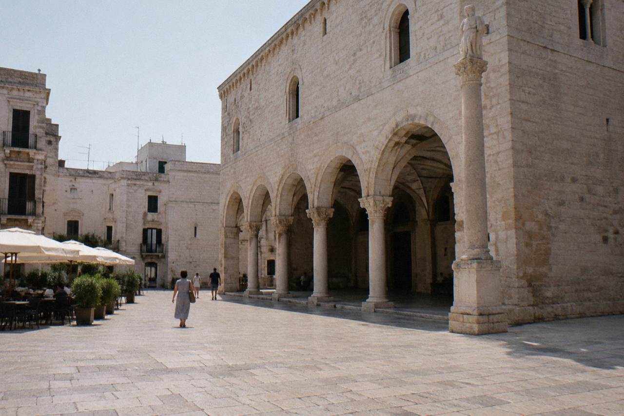 Old town square with a medieval arcaded loggia, a slender stone column and cafe tables under parasols in soft morning light
