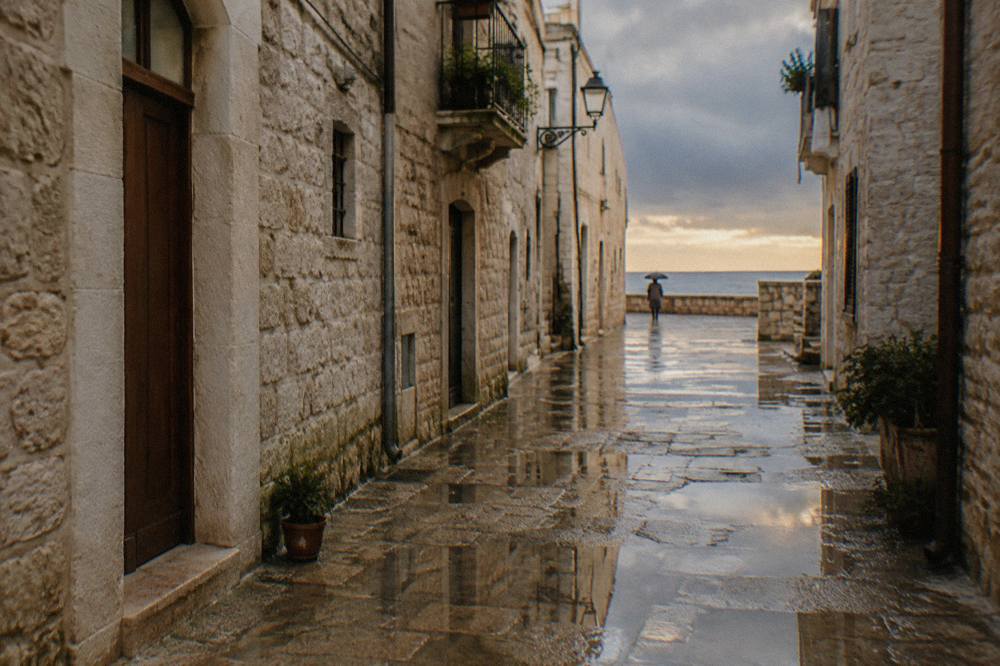 Wet limestone old town lane after rain with reflections in puddles and a distant person under an umbrella