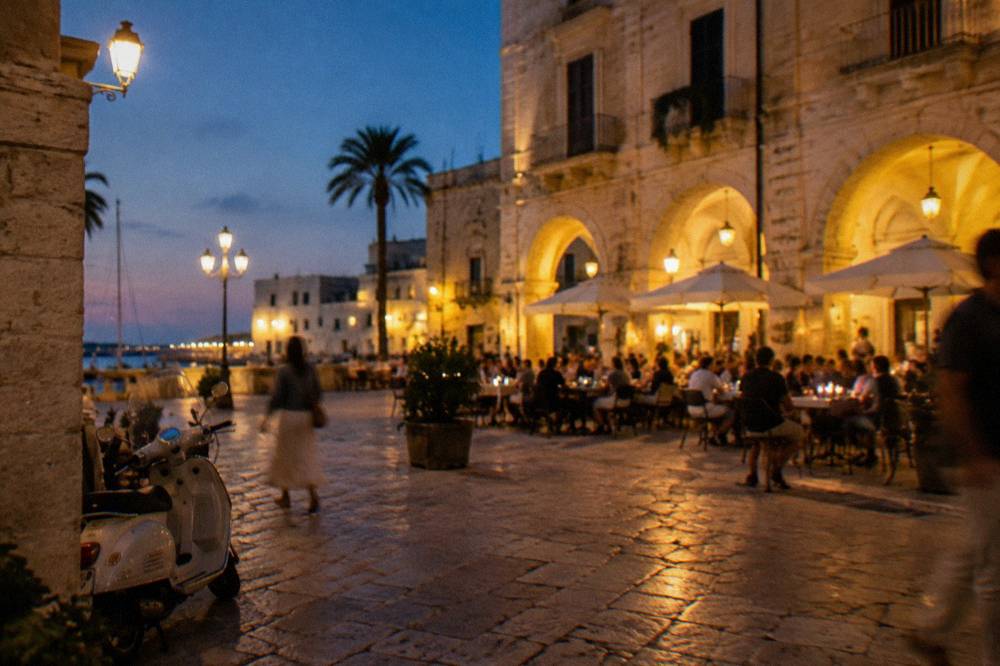 Evening piazza with warm light on cream walls, out-of-focus diners at tables and glowing market arches