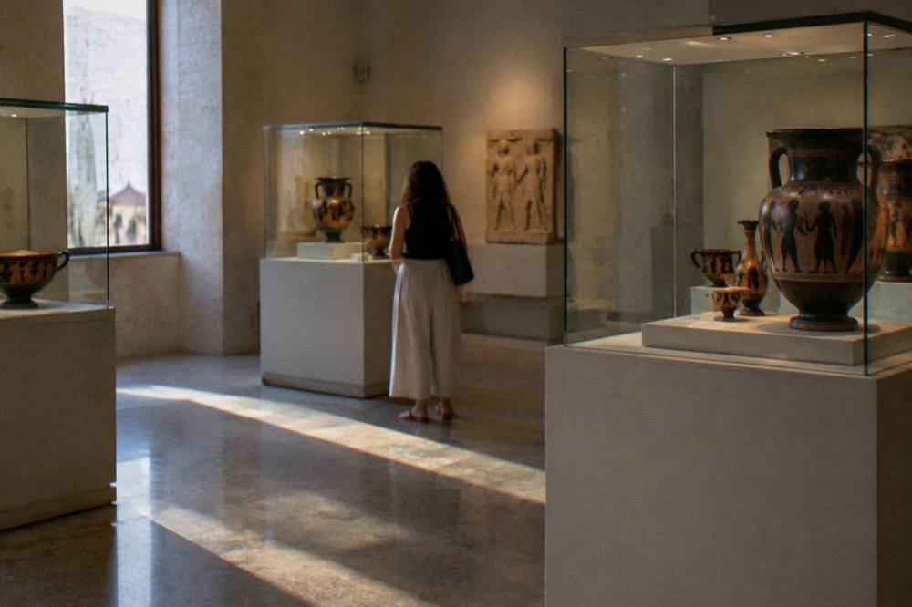 Quiet museum room with glass cases of ancient painted pottery under soft spotlighting and one visitor from behind