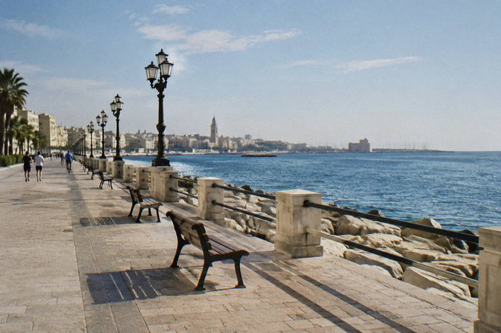 Wide paved seafront promenade with distant joggers and cyclists, benches and calm blue sea in late afternoon