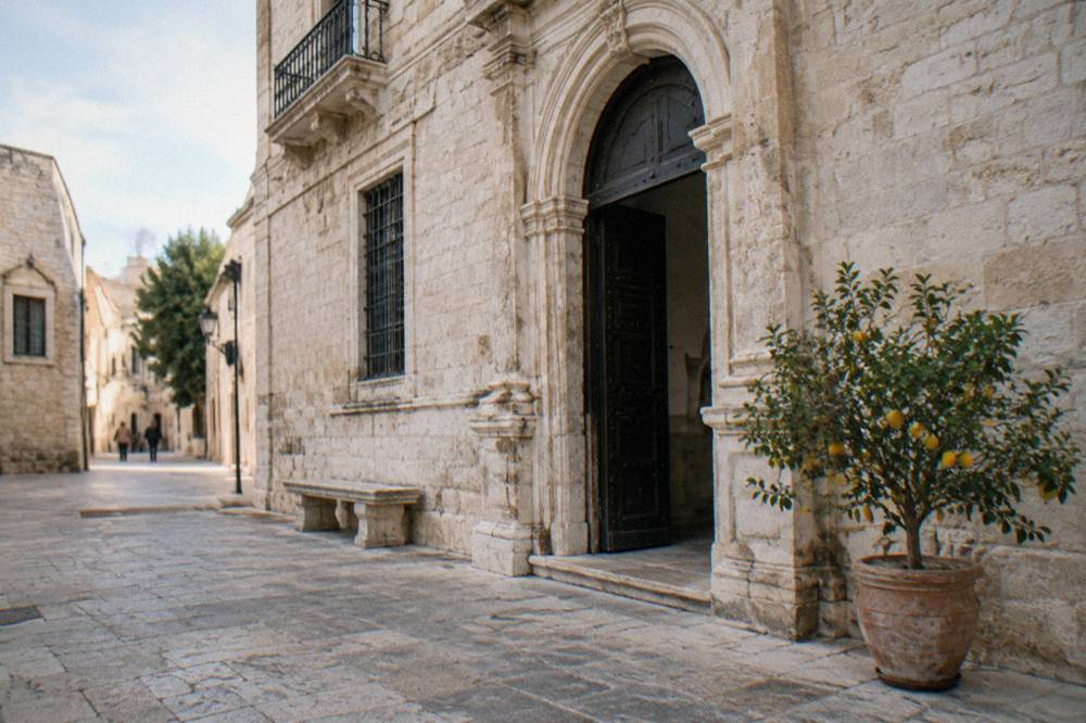 Pale stone palazzo with an arched portal on a quiet old-town street beside a stone bench and a potted lemon tree