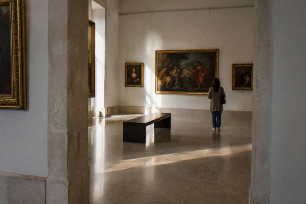 Gallery room with framed old paintings on pale walls, a central bench and one visitor seen from behind