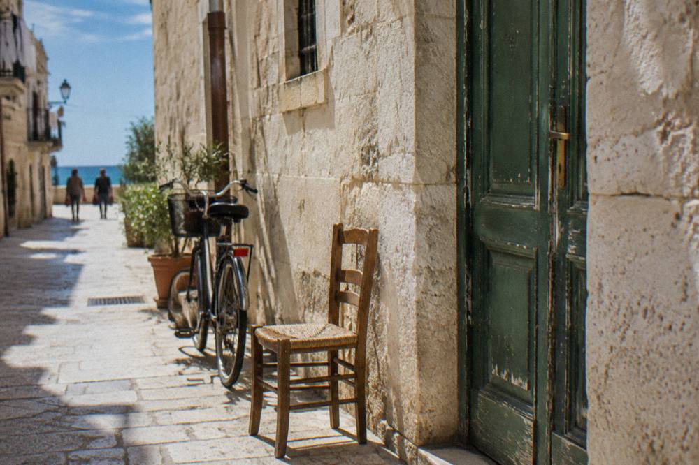 Faded green wooden door in a pale limestone wall with a chair and a bicycle beside it