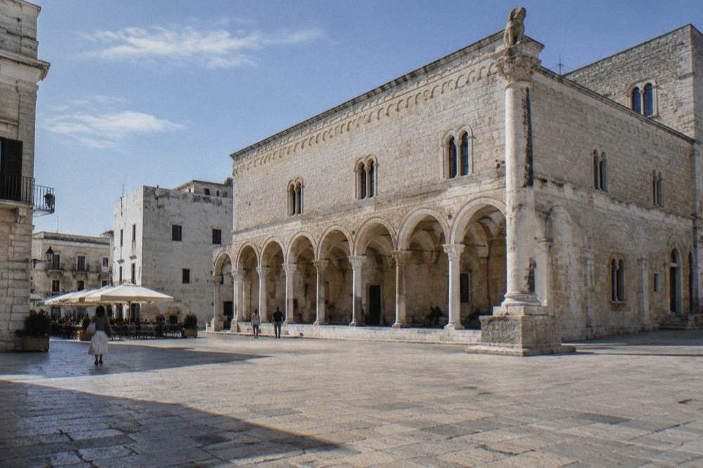 Old town square with a medieval arcaded loggia, a slender stone column and cafe tables under parasols in soft morning light