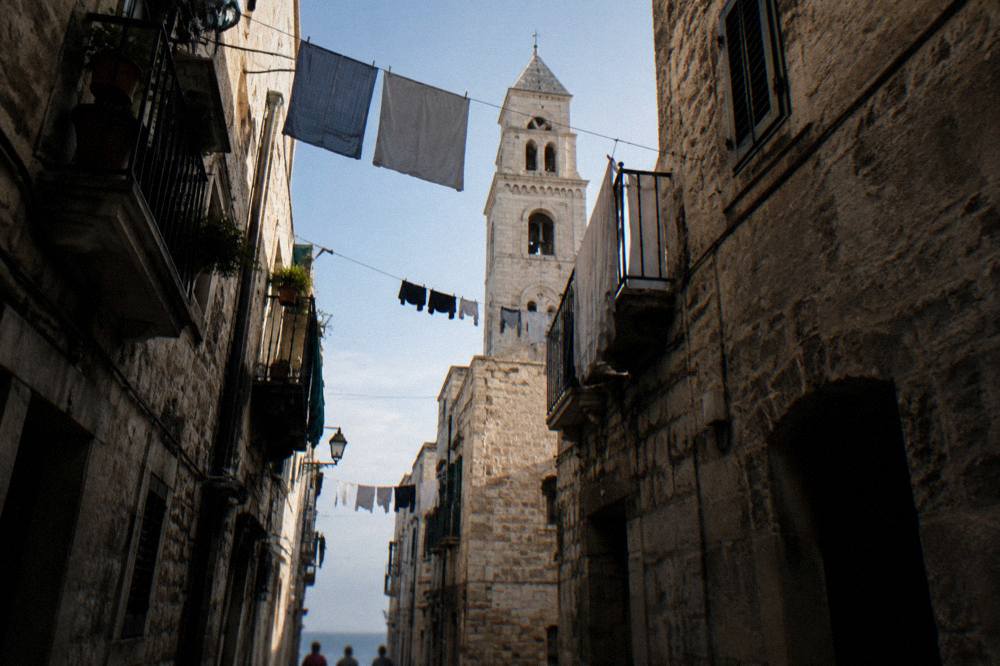 Looking up between close walls at a strip of blue sky crossed by washing lines and a bell tower