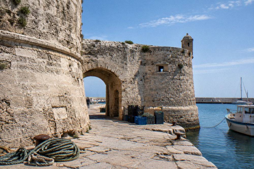 Weathered limestone fort walls and a low arched gateway beside harbour water with coiled ropes and crates on the quay