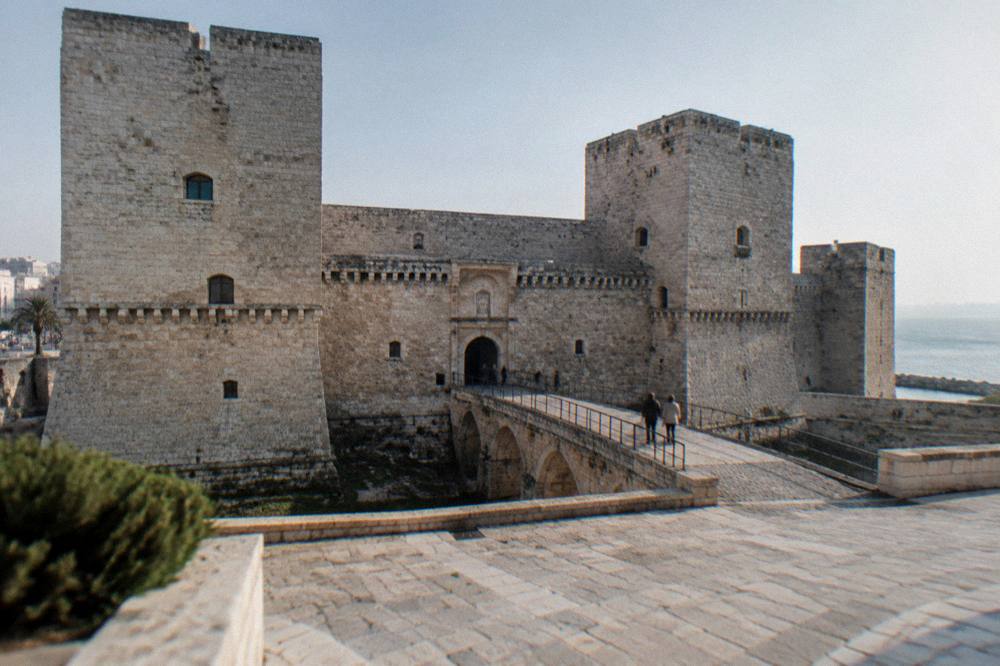 Massive square pale limestone fortress with corner towers, a dry moat and a bridge leading to an arched gate