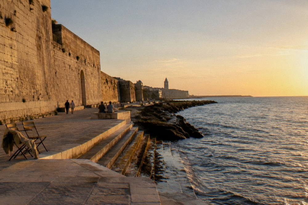 Massive pale limestone sea wall with a promenade at its foot and worn steps down to the water in low afternoon sun