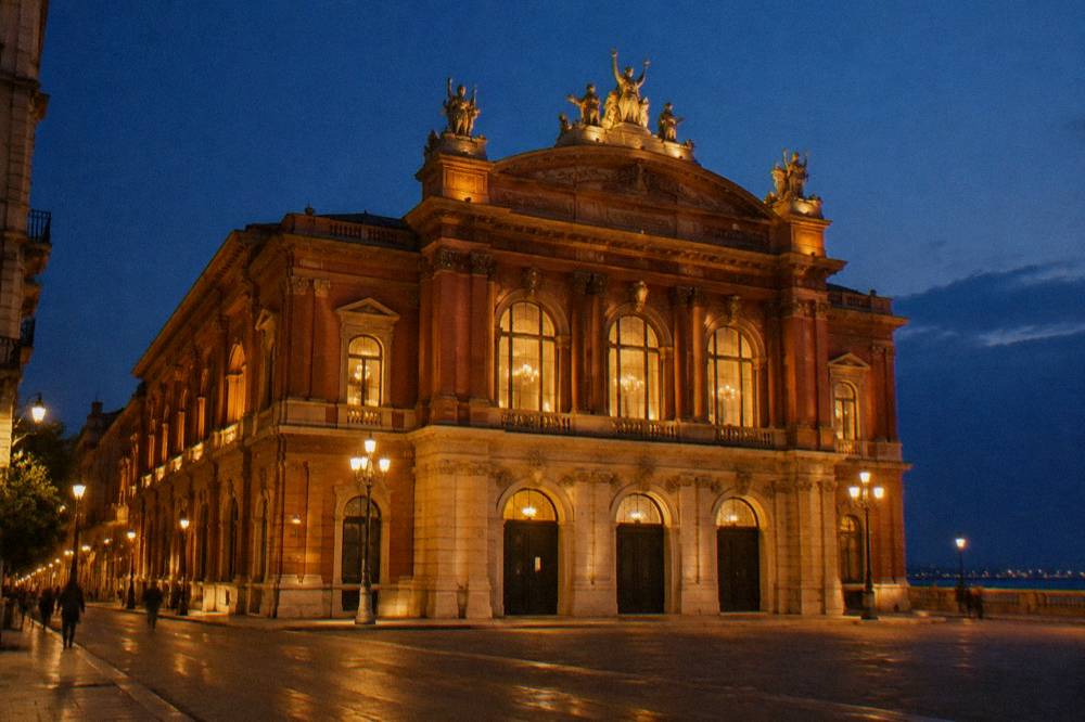 Neoclassical theatre facade floodlit at evening with glowing arched windows and a deep blue sky
