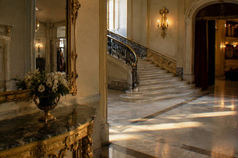 Quiet gilded theatre foyer with a marble staircase and a tall mirror in soft warm light