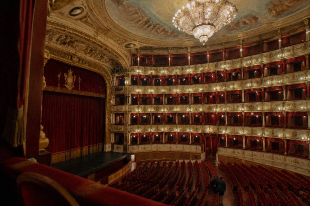 Empty horseshoe-shaped theatre with tiers of red and gold boxes, a large chandelier and a painted ceiling