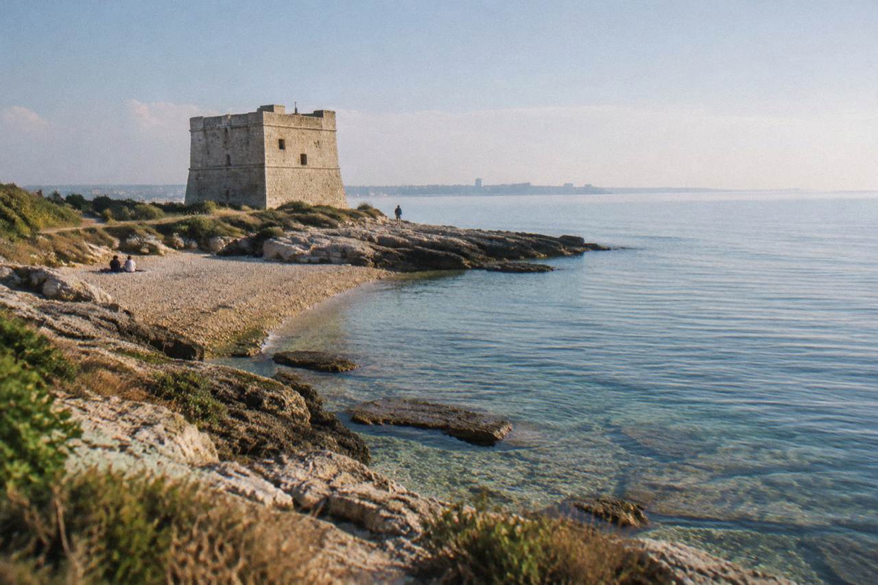 Squat square stone watchtower on a flat rocky shore beside a small pebble beach and low scrub