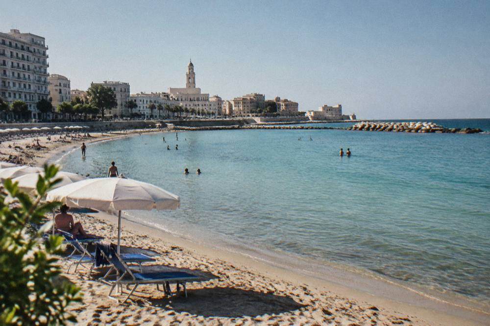Small urban sandy beach with parasols in a curved bay, shallow turquoise water and seafront buildings behind