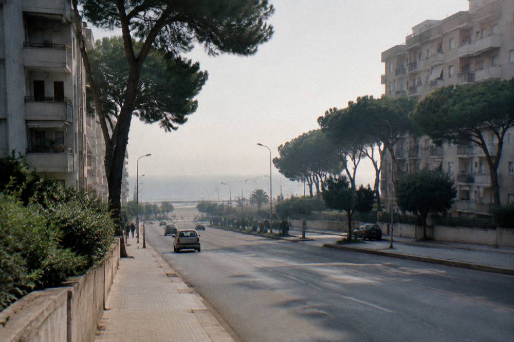 Residential district of 1960s apartment blocks with a wide road leading to a strip of sea between pine trees