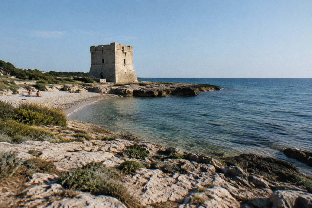 Squat square stone watchtower on a flat rocky shore beside a small pebble beach and low scrub