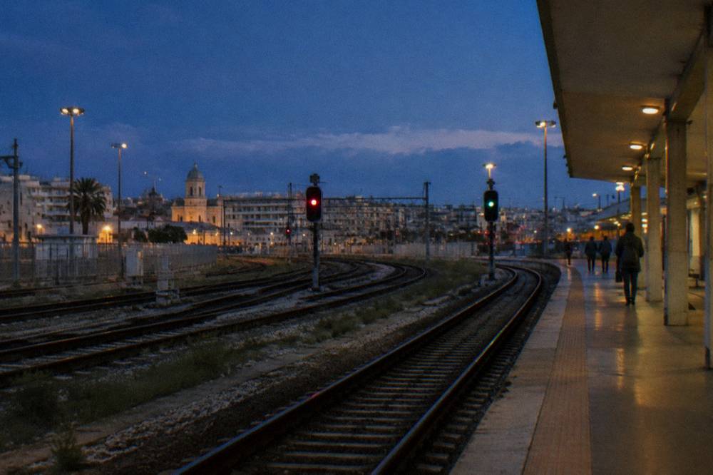 Rails curving away from a platform end at dusk with glowing signal lights and a few distant passengers