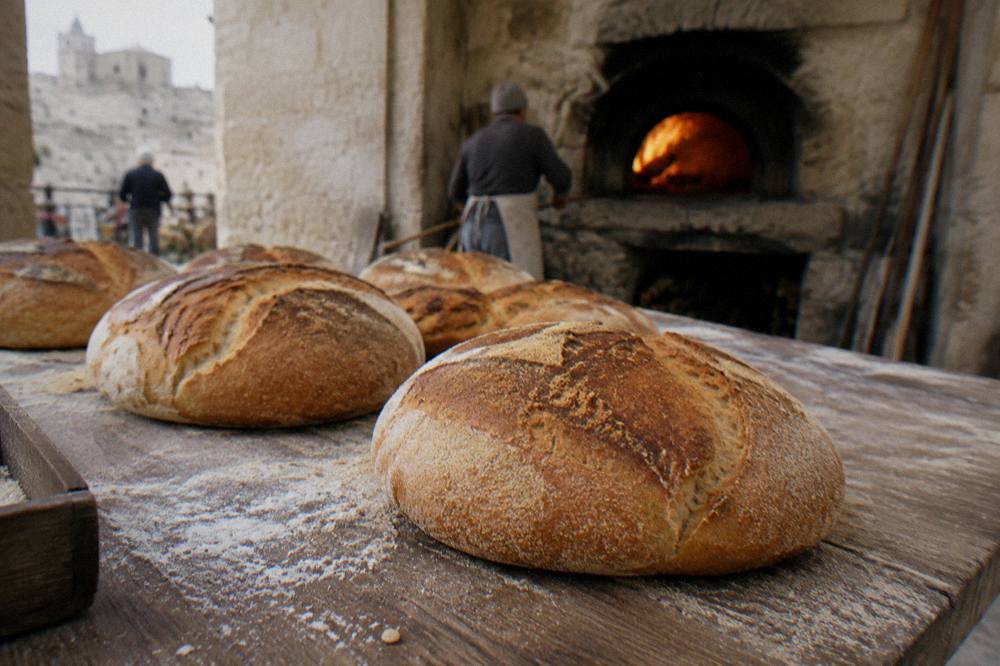 Large round loaves of semolina bread with thick golden crusts on a wooden table beside a wood-fired oven