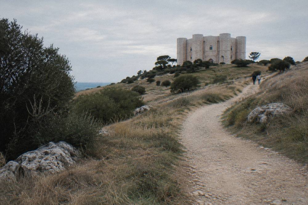 Dusty path winding up a hill through dry grass towards a distant pale octagonal castle in late afternoon