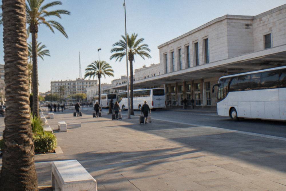 Station forecourt with plain white buses, palm trees and distant travellers with wheeled suitcases