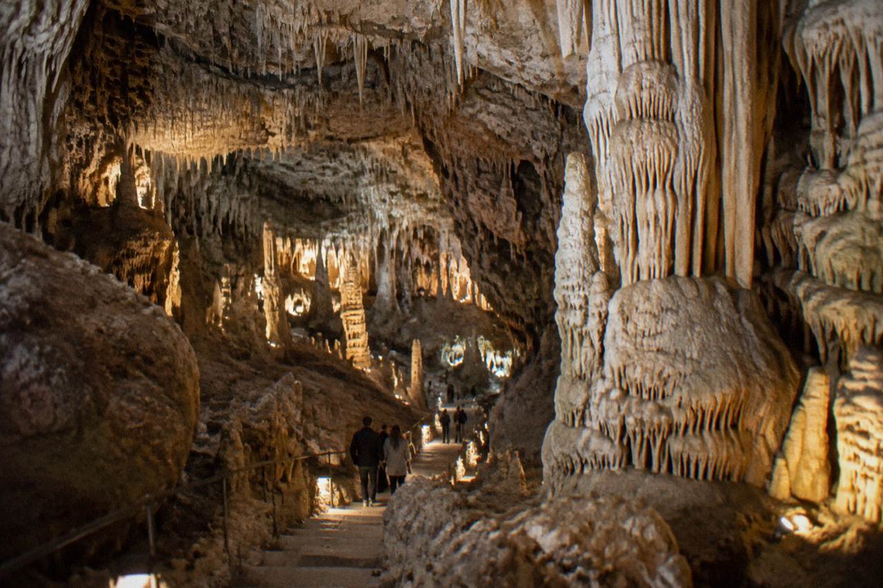 Vast underground cave hall with pale stalactites and stalagmites under warm lights and tiny visitors on a stepped path