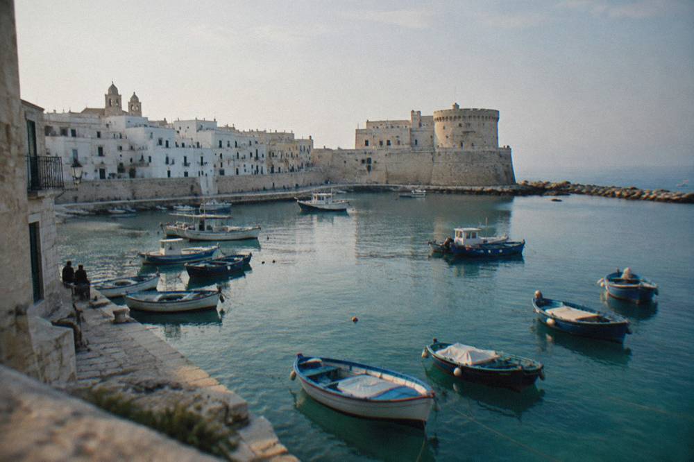 Old harbour of small painted wooden boats with a stone castle on the headland and a whitewashed old town behind