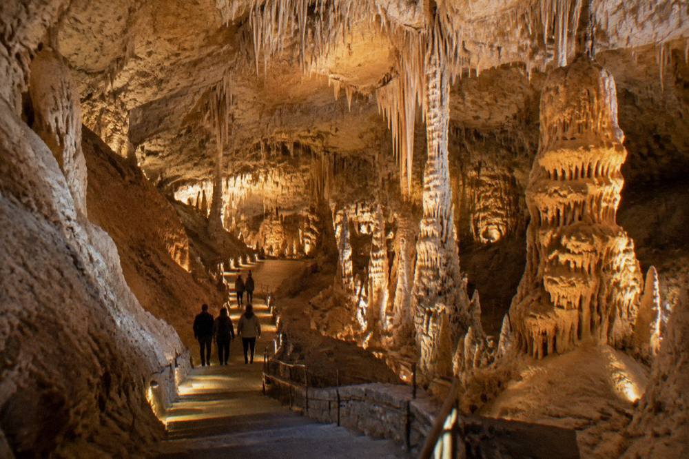 Vast underground cave hall with pale stalactites and stalagmites under warm lights and tiny visitors on a stepped path