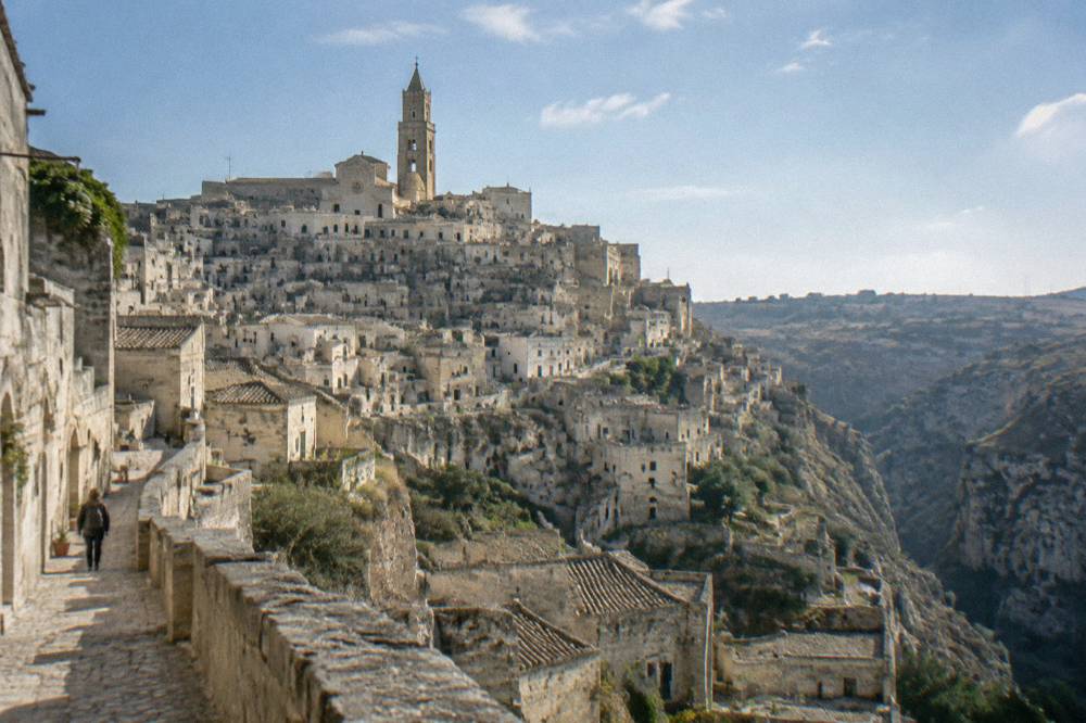 Tiers of honey-coloured cave dwellings stacked on the side of a deep ravine below a cathedral bell tower
