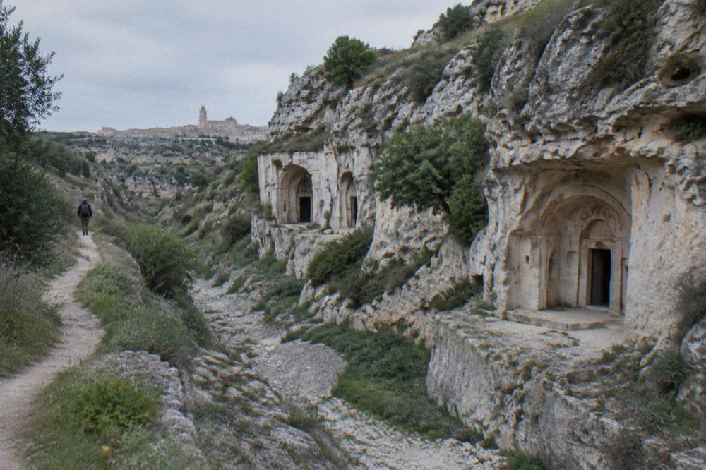 Cave entrances cut into a pale rock face above a dry stream bed with shrubs on the ledges and a distant hiker