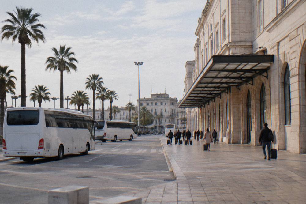 Station forecourt with plain white buses, palm trees and distant travellers with wheeled suitcases