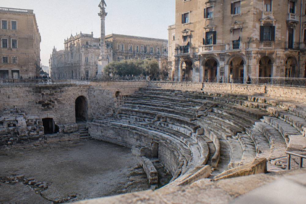 Roman amphitheatre half excavated in a city square surrounded by palazzi in afternoon light