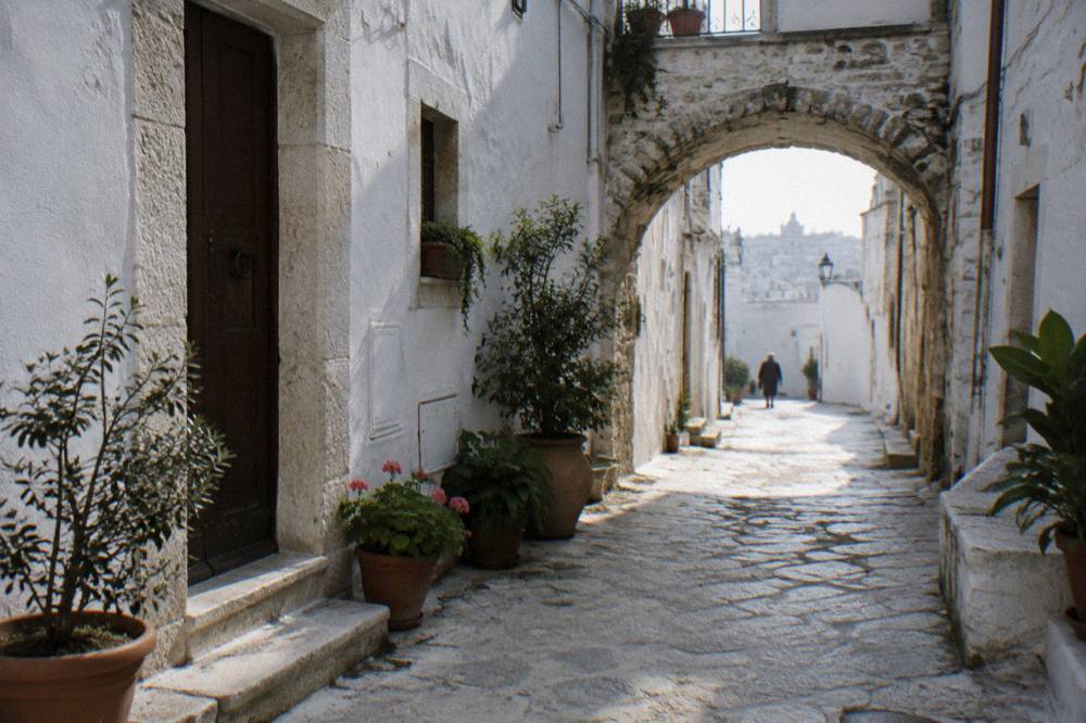 Narrow white old town lane with a stone arch and potted plants in afternoon shade