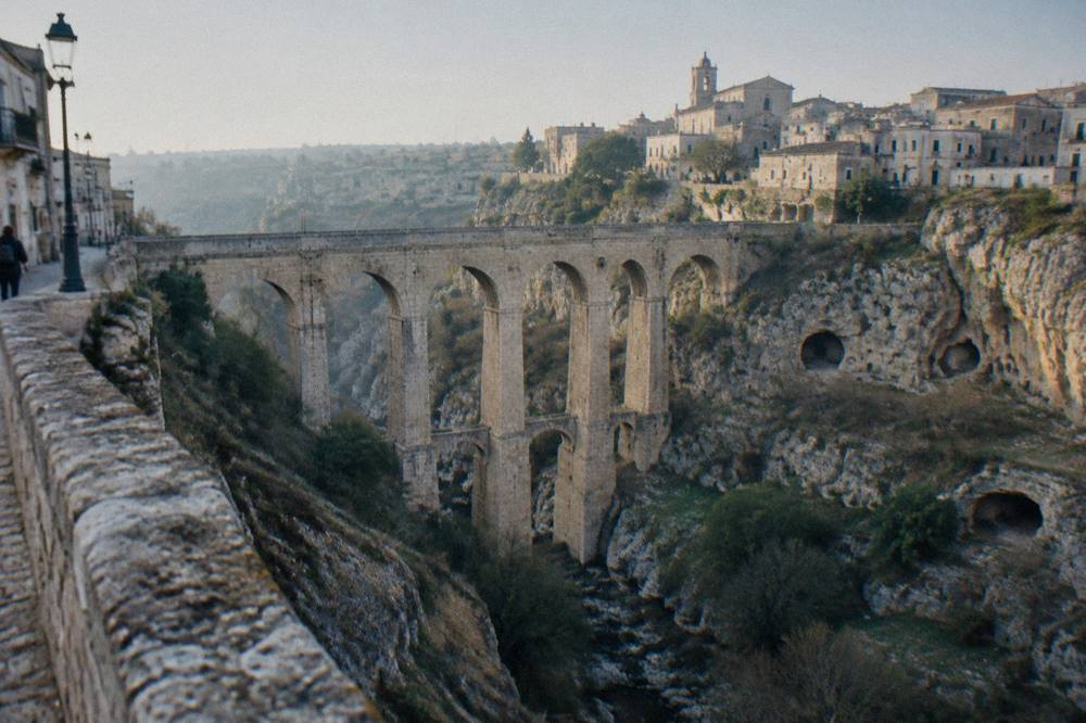 Deep rocky ravine crossed by a tall many-arched stone bridge with old town houses on the far rim