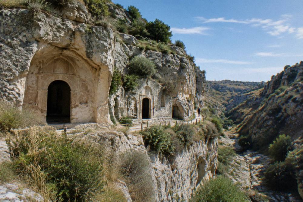 Cave entrances cut into a pale rock face above a dry stream bed with shrubs on the ledges and a distant hiker