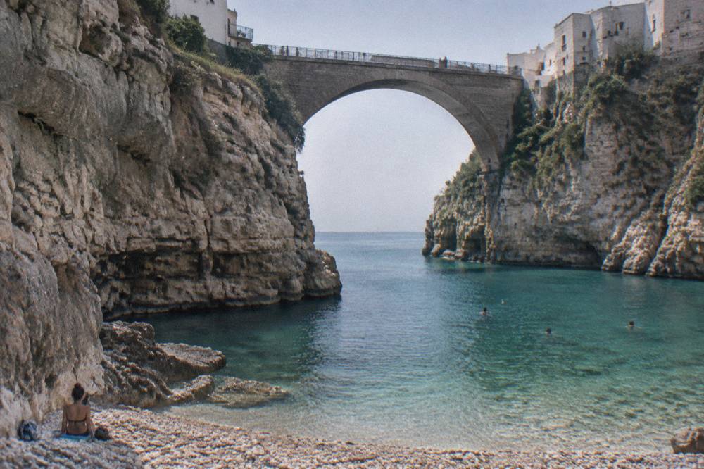 Narrow pebble cove between steep limestone cliffs under a stone arch bridge with turquoise water