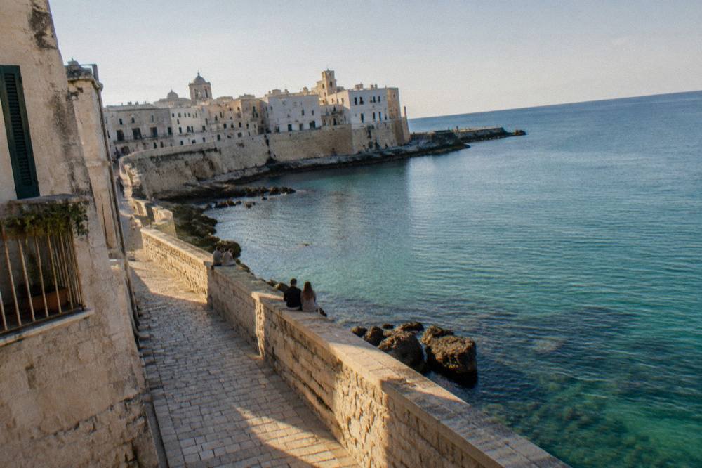 Sea promenade along an old town wall above calm turquoise water with distant people sitting on the wall